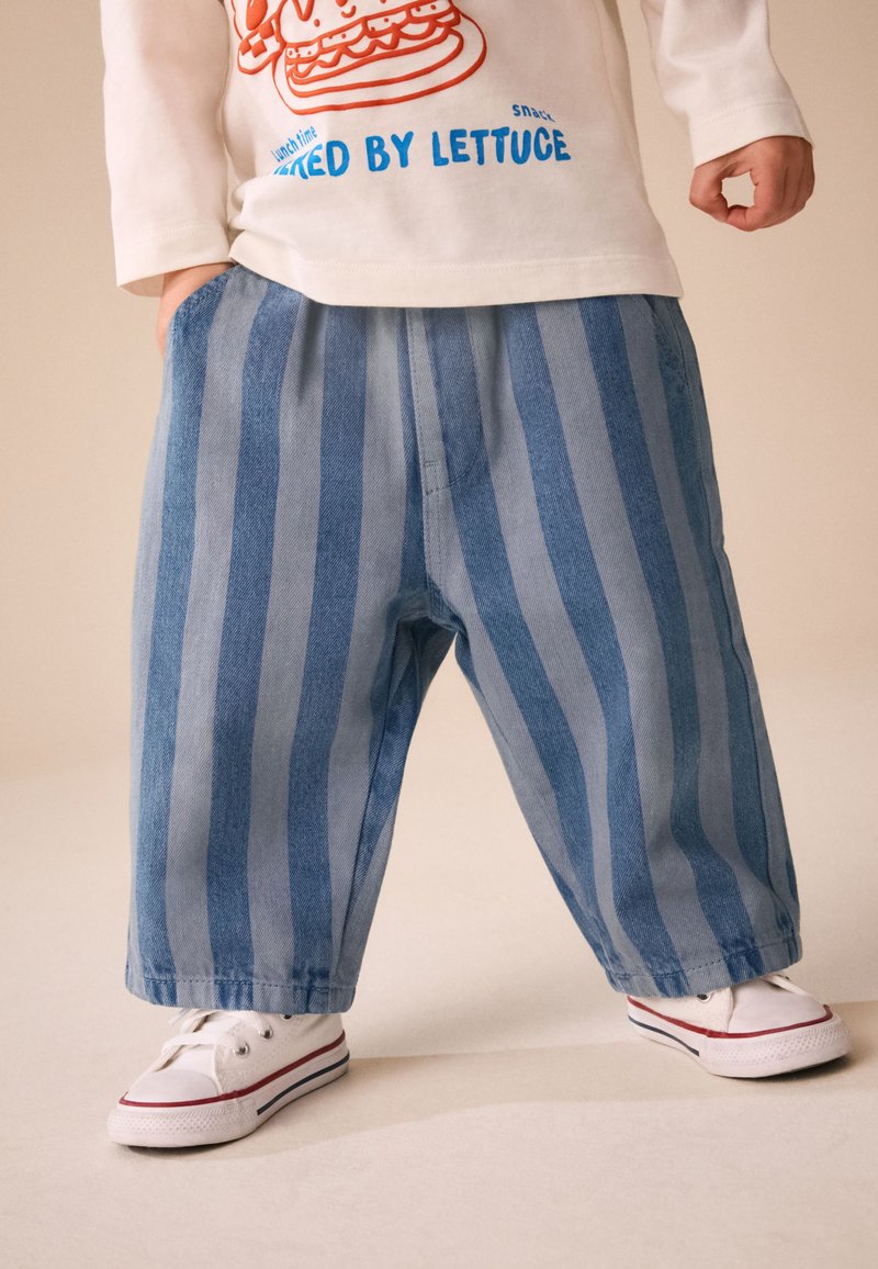 Niño con pantalones anchos de rayas verticales azul y azul claro, zapatillas blancas y una camiseta de manga larga con un estampado de sándwich.
