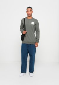 Grå langermet hettegenser med en lapp-logo, sammen med løse blå denimbukser. Svart crossbody-veske, hvite sneakers. Enkel bakgrunn.