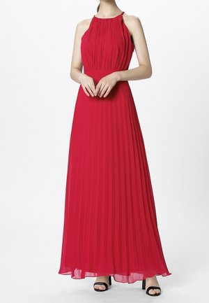Femme portant une robe rouge plissée longue au sol, col halter, avec des sandales noires à talons hauts ouvertes à l’avant, debout devant un fond blanc.
