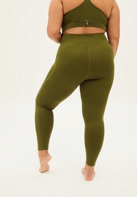 Osoba plus-size w oliwkowozielonych legginsach z wysokim stanem i pasującym stanikiem sportowym, stojąca boso na jasnym tle.