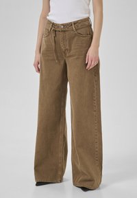 Pantalones de pana marrón de pierna ancha con cintura alta, cinturón ajustable, bolsillos delanteros y detalles de costura visibles.