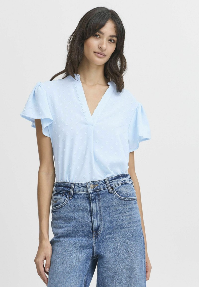 Blusa azzurro chiaro a maniche corte con scollatura a V, caratterizzata da un pattern a pois testurizzati. Abbinata a jeans in denim a vita alta.
