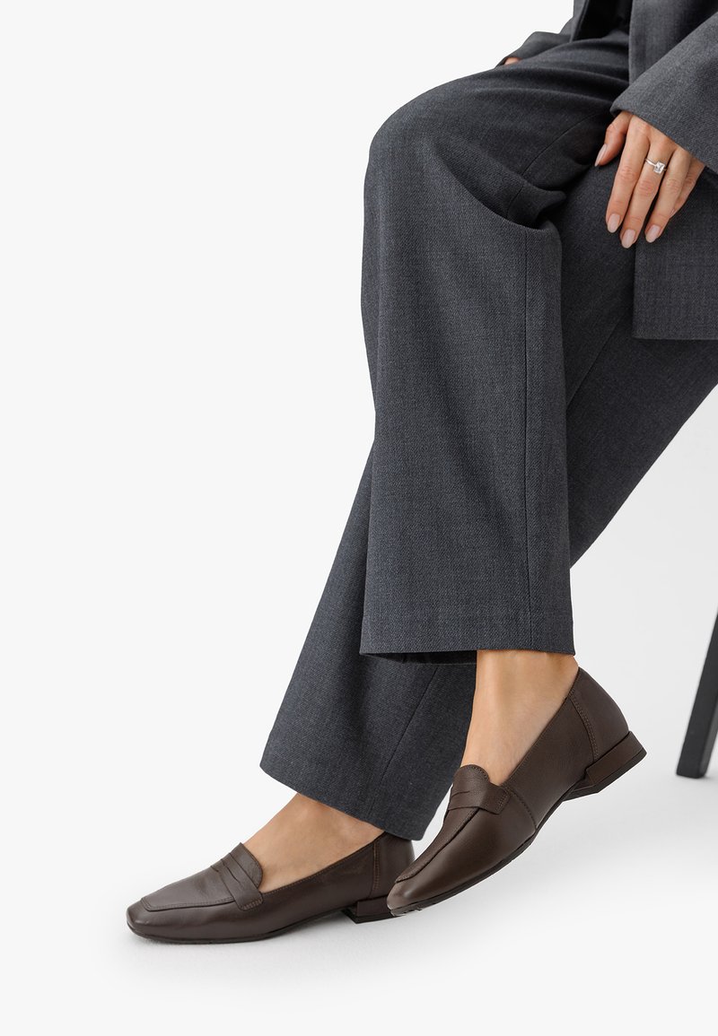 Pruunid nahast loafers, millel on ümar varvas, õmmeldud rihma detail ja lame konts, kantud hallide kohandatud pükstega.