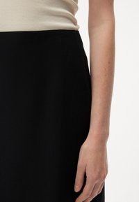 Czarna spódnica z gładkiego materiału, charakteryzująca się prostym wzorem. Zbliżenie na dłoń opierającą się na tkaninie spódnicy, ukazujące teksturę.