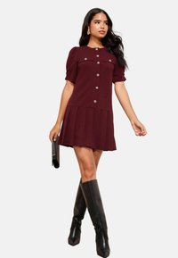 Rochie burgundă cu mâneci scurte bufante, nasturi în față și fustă plisată. Asortată cu cizme negre până la genunchi și plic negru texturat.