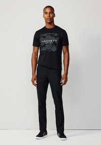 Camiseta negra con gráfico de "HACKETT SPORT", combinada con pantalones negros y zapatillas negras. Textura suave, diseño ajustado. Look simple y monocromático.