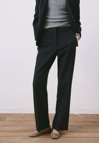 Persona vestida con pantalones oscuros de rayas finas, top gris acanalado, blazer oscuro y zapatos planos beige, de pie sobre un suelo de madera contra una pared blanca.