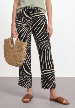 Personne portant un pantalon large à motifs noir et beige, un haut vert olive, des sandales marron et tenant un sac en paille tissé.
