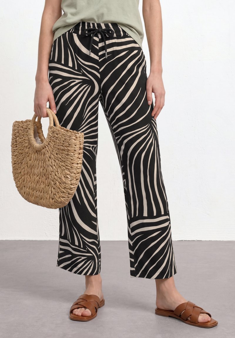 Personne portant un pantalon large à motifs noir et beige, un haut vert olive, des sandales marron et tenant un sac en paille tissé.