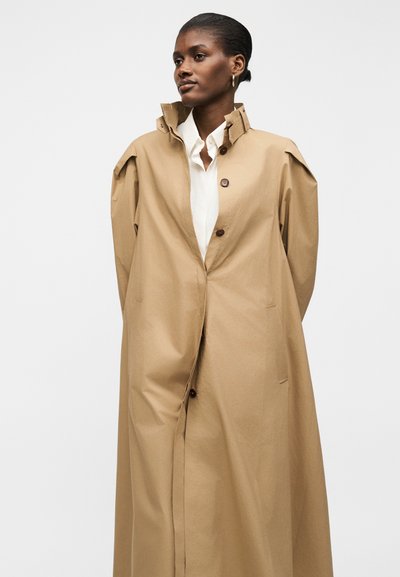 Femme portant un long trench-coat beige par-dessus une chemise blanche à col, debout les mains dans le dos, regardant sur le côté sur un fond uni.