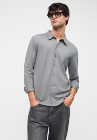 Γκρι πουκάμισο με κουμπιά και μακριά μανίκια, μαλακή υφή και κλασικό γιακά. Φορεμένο με σκούρα γκρι τζιν, τα χέρια στις τσέπες.