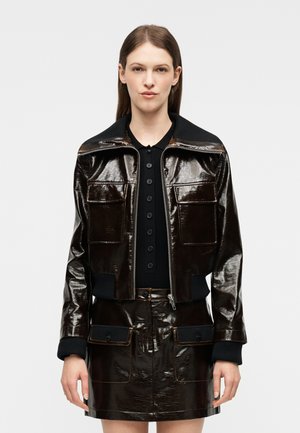 Vrouw die een glanzend donkerbruine jas met rits draagt, voorzien van grote zakken aan de voorkant, en een bijpassende rok met klepzakken, staand tegen een effen achtergrond.