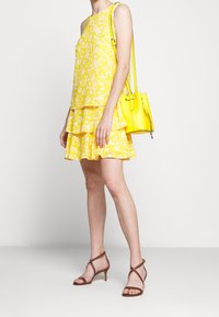 Žlutý květinový bezrukávový top vrstvený přes odpovídající sukni s volány; doplněný žlutou kabelkou a hnědými páskovými podpatky.