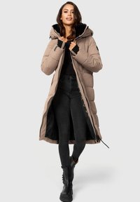 Abrigo de plumas largo en color beige con capucha forrada en negro, cierres de cremallera y a presión, bolsillos laterales y textura acolchada. Lleva puesto sobre pantalones negros y botas.