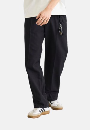 Person iført løse sorte bukser med nøglering på bæltestrop og hvide sneakers med sorte striber og gummi såler.