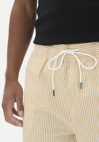 Vyrų torsas su tamsia oda, vilkintis juodą beprotišką marškinėlį ir šviesiai bei baltais dryžiais išmatuotus šortus, su balta virvele.