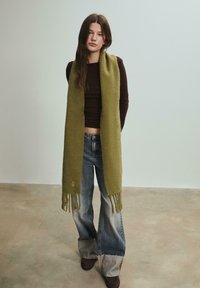 Écharpe vert olive en matériau doux, avec des bords à franges. Portée par-dessus un haut brun ajusté et associée à un jean en denim évasé.