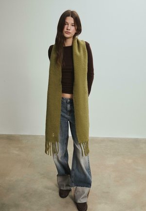 Olivově zelený šátek vyrobený z měkkého materiálu, s třásněmi na okrajích. Nositý přes přiléhavý hnědý top a kombinovaný s rozšířenými džínovými kalhotami.