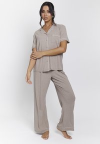 Conjunto de pijama marrón claro de manga corta con ribete blanco. Presenta una camisa abotonada con un bolsillo y pantalones anchos y relajados. Textura de tela suave.