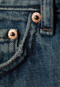 Denim jeans detail featuring dark blue fabric, copper rivets, and contrasting stitching on the pockets and waistband. Textured surface visible.