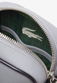 Interno della borsa grigio testurizzato con motivo a griglia verde. Presenta cerniere argento e un logo in pelle di coccodrillo bianco. Include una tasca con zip separata.