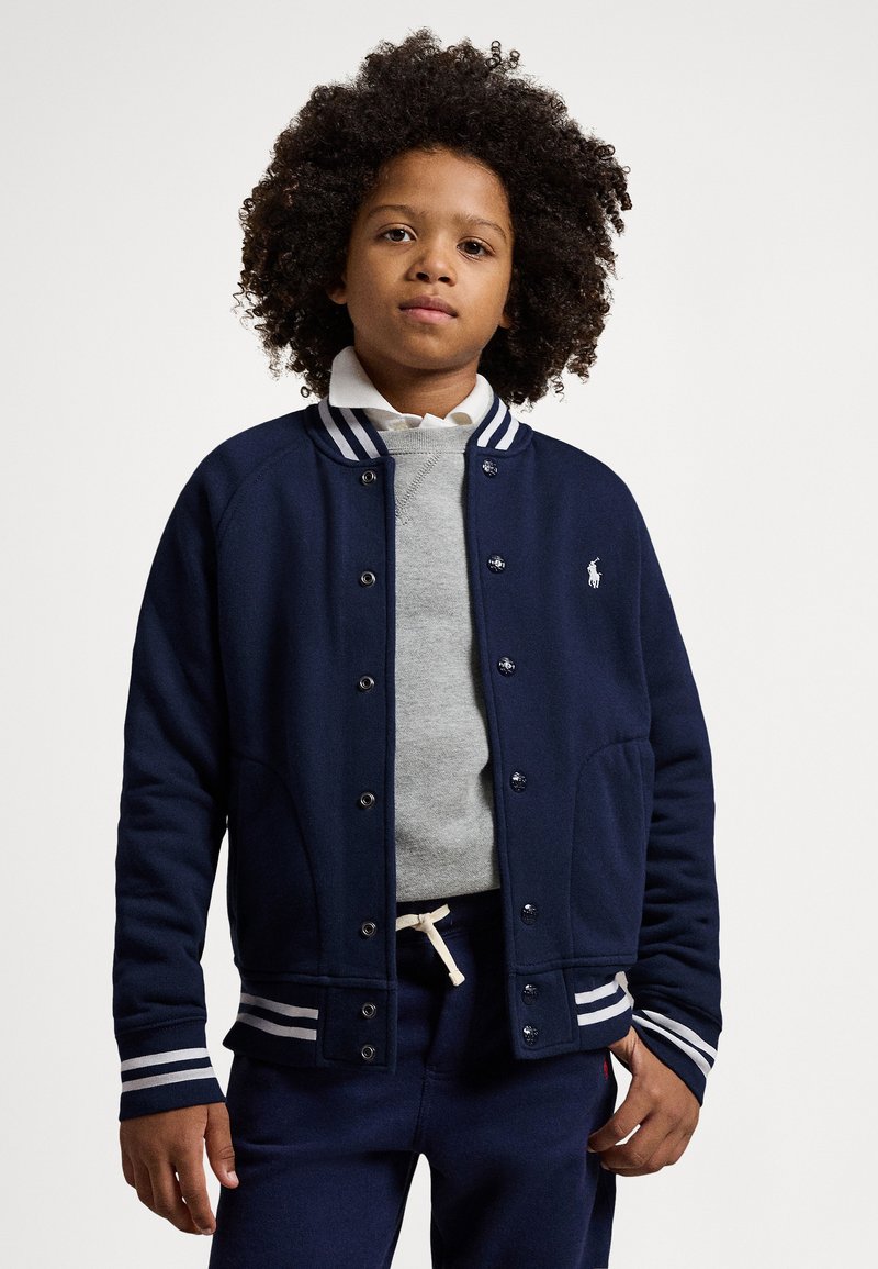 Marinblå varsityjacka med vitrandig krage och mudd, har tryckknappar, matchad med en grå tröja och mörka byxor.