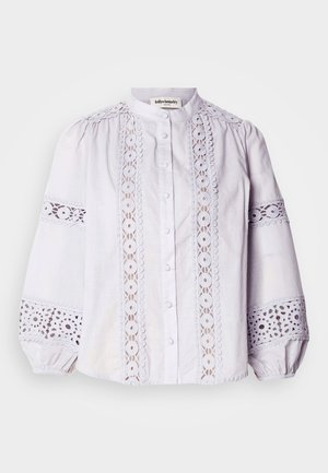 Balta blūze ar augstu apkakli, pogu aizdari un pūkainām piedurknēm. Iezīmējas sarežģītiem mežģīņu paneļiem ar apļveida rakstiem pa priekšpusi un piedurkņu malām.