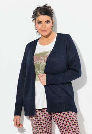 Marineblauer Strickcardigan mit offenem Fall, strukturiertem Stoff und gerippten Bündchen, über einem weißen T-Shirt mit grünem Grafikdesign und gemusterten rosa Hosen.