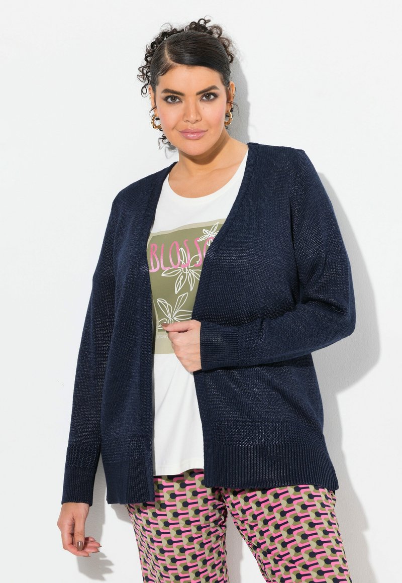 Marineblauer Strickcardigan mit offenem Fall, strukturiertem Stoff und gerippten Bündchen, über einem weißen T-Shirt mit grünem Grafikdesign und gemusterten rosa Hosen.