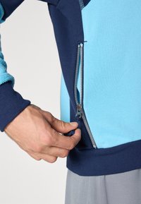 Sweatshirt com fecho éclair, apresentando blocos de cor azul e marinho, mangas texturizadas e um fecho éclair prateado visível com aba.