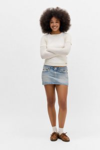 Leende kvinna med afrohår som bär en krämfärgad långärmad topp, denim-minikjol, vita strumpor och bruna mockaskor, med armarna korsade.