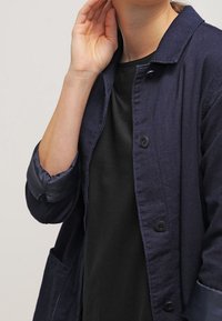 Mörkblå, texturerad jacka med krage, knappstängning och stora fickor. Uppvikta ärmar har kontrasterande tyg. Svart topp under.