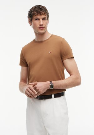 Homme aux cheveux bouclés portant un t-shirt marron à manches courtes, un pantalon blanc, une ceinture noire et une montre-bracelet, debout devant un fond blanc.