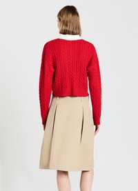 Roter Kabelstrickpullover mit langen Ärmeln, kombiniert mit einem beigefarbenen Faltenrock. Der Pullover hat gerippte Bündchen und einen gerippten Saum, was einen Kontrast in der Textur schafft.