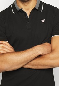 Czarna koszulka polo z kołnierzykiem, z szarym paskiem dekoracyjnym. Zawiera trzy guziki oraz małe trójkątne logo na lewej piersi.