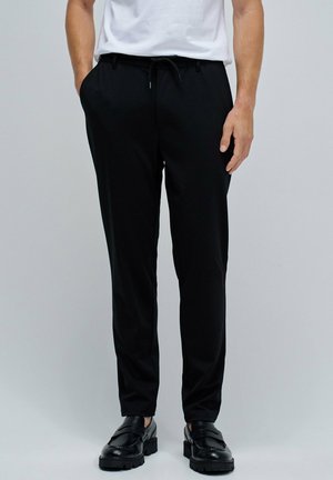 Hombre que lleva pantalones negros con cordón, mocasines negros y una camiseta blanca, de pie con una mano en el bolsillo contra un fondo liso.