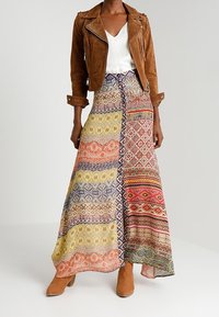 Bruine suède bikerjack over een wit shirt, gecombineerd met een multi-gekleurde maxi-rok met geometrische en bloemmotieven. Bruine enkellaarsjes.