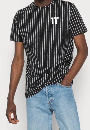 Mann trägt ein schwarzes T-Shirt mit weißen vertikalen Streifen und dem "11°"-Logo, kombiniert mit hellblauen Jeans, die Hände in den Taschen.