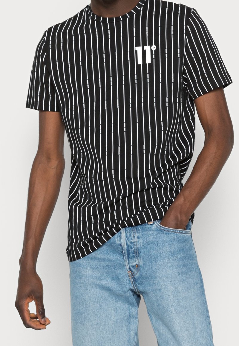 Homme portant un t-shirt noir avec des rayures verticales blanches et le logo "11°", associé à un jean en denim bleu clair, la main dans la poche.