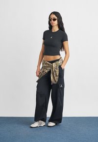 Μαύρο κοντομάνικο μπλουζάκι με κεντημένο λογότυπο, πουκάμισο με καμουφλαρισμένο μοτίβο δεμένο στη μέση, φαρδιά μαύρη τζιν παντελόνια cargo και λευκά αθλητικά παπούτσια με μαύρες λεπτομέρειες.