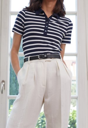 Vrouw draagt een navyblauw-wit gestreept poloshirt, ingestopt in een high-waist crème broek met zwarte riem, staand binnen naast een raam.