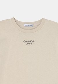 Béžové bavlněné tričko s kulatým výstřihem, krátkým rukávem a černým potiskem "Calvin Klein Jeans" na oblasti hrudi.