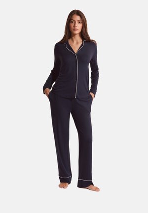 Femme debout, pieds nus, portant un haut de pyjama bleu marine à manches longues et boutonné, ainsi qu'un pantalon assorti avec des détails en piping blanc.