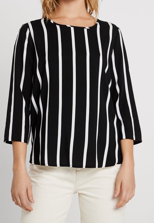 Kvinna iklädd en svart långärmad tröja med vertikala vita ränder och ljusa byxor mot en enkel bakgrund.