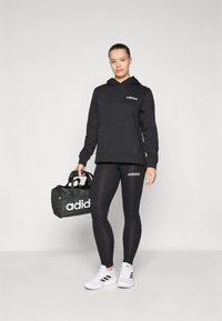 Svart hoodie med framficka och vit logotyp, kombinerad med svarta leggings med logotyp på sidan. Håller en svart gymväska. Vita sneakers.
