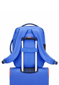 Sac à dos bleu fixé à la poignée d'une valise bleue avec poignée argentée rétractable et accents rouges aux coins sur fond blanc.