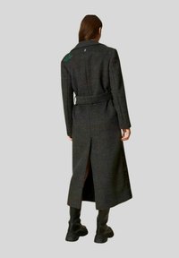Cappotto lungo a quadri neri e grigi con vita regolabile, schiena spaccata e piccolo dettaglio ricamato sulla spalla. Texture in lana, design sartoriale.