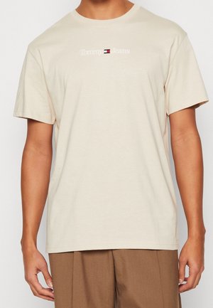 T-shirt en coton beige avec des manches courtes et un col rond, affichant le texte du logo "Tommy Jeans" et un petit emblème de drapeau au centre.