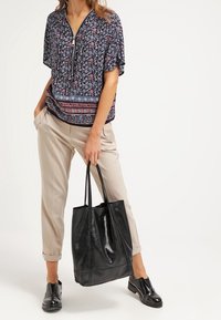 Nainen, jolla on päällään kukkakuosiset puserot ja vaaleanbeiget 3/4-housut, pitää kädessään suurta mustaa nahkakassia ja on pukeutunut mustiin loafereihin.