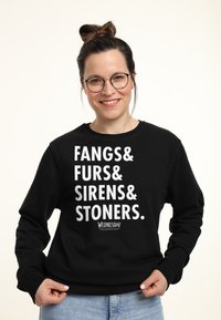 Persona sorridente con occhiali che indossa un felpa nera con la scritta "Fangs & Furs & Sirens & Stoners" in bianco, mani che afferrano l'orlo.